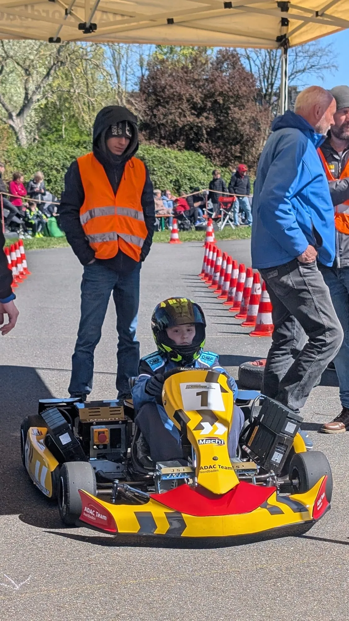 Nachwuchsfahrer im gelben ADAC-Kart mit der Startnummer 1 im Vorstartbereich, Streckenposten daneben