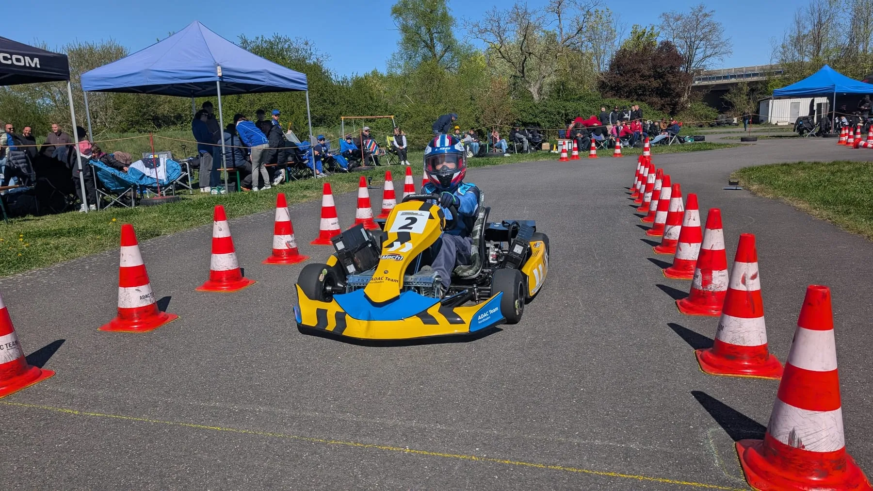 Gelb-blaues ADAC-Elektrokart im Pylonenparcours des Vereinsplatzes, Zuschauer unter Pavillons am Rand