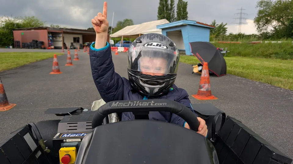 Kind mit Helm im Kart hebt den Zeigefinger, im Hintergrund Pylonen und die Vereinsräumlichkeiten