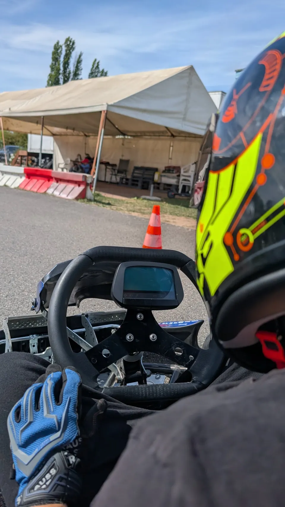 Fahrer mit gelb-schwarzem Helm und blauen Handschuhen am Lenkrad, dahinter das Fahrerlager