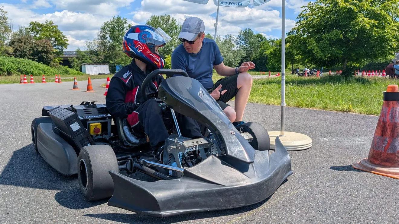 Übungsleiter kniet neben einem Kind im Kart und erklärt die Bedienung, Pylonen im Hintergrund