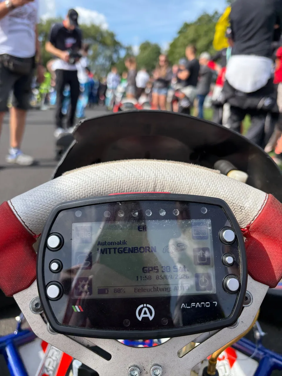Blick aus dem Kart auf das Lenkrad-Display, im Hintergrund das Fahrerlager